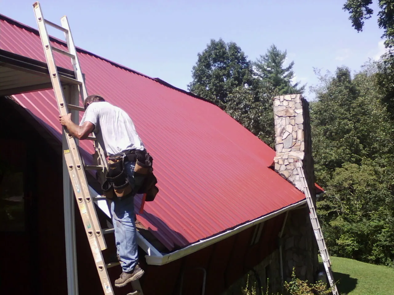 Roofer on a ladder beside a red metal roof and stone chimney — Soffit & Fascia Repair by Roofing Brothers in Morristown
